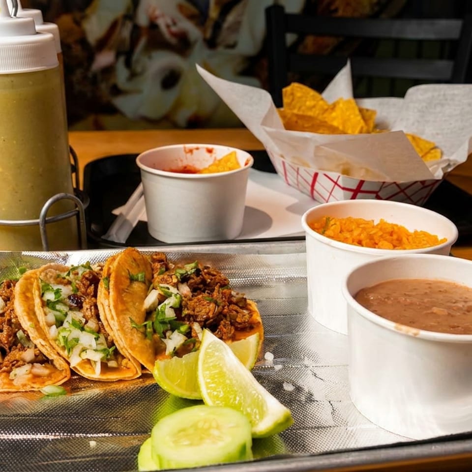 Tray of three tacos topped with onions and cilantro, lime wedges, beans, rice, chips, and salsa on a restaurant table.